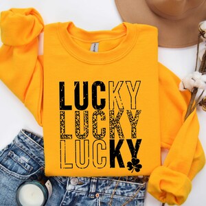 May include: A yellow sweatshirt with the words "LUCKY" printed three times in black. The second "LUCKY" is printed in a leopard print pattern. A small shamrock is printed below the third "LUCKY".