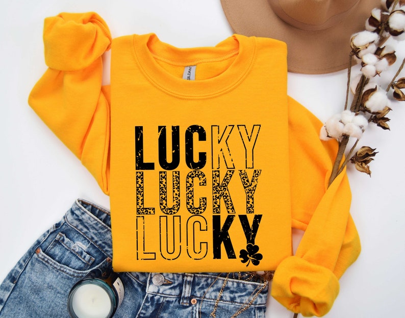May include: A yellow sweatshirt with the words "LUCKY" printed three times in black. The second "LUCKY" is printed in a leopard print pattern. A small shamrock is printed below the third "LUCKY".