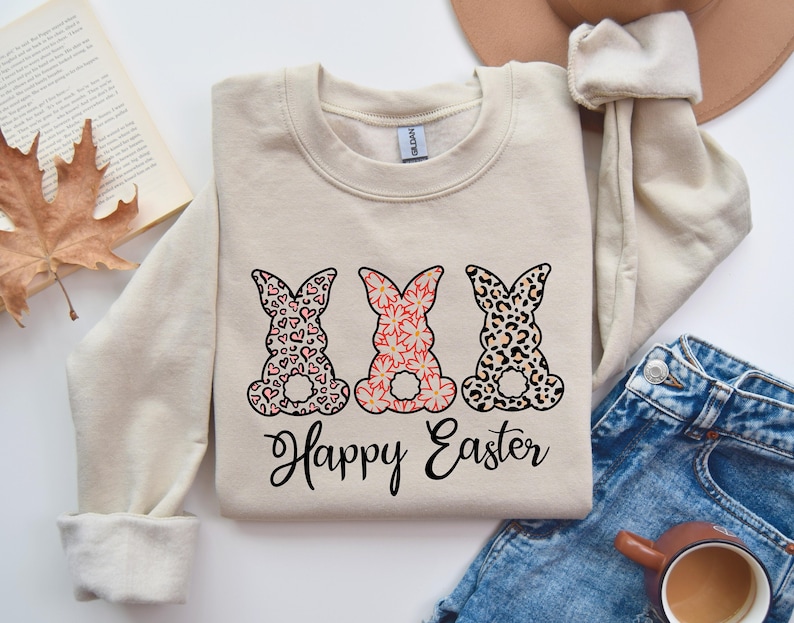 Puede incluir: Una sudadera beige con tres siluetas de conejos en diferentes patrones: corazones rosas, flores rosas y estampado de leopardo. Los conejos est&aacute;n alineados en una fila y el texto "Happy Easter" est&aacute; impreso debajo.
