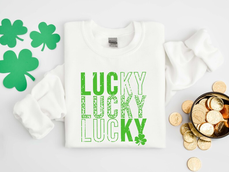 May include: White sweatshirt with the words "LUCKY" repeated three times in green text with a distressed look. The letters in the second "LUCKY" are filled with green shamrocks. A small green shamrock is in the bottom right corner.