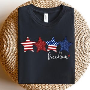 May include: Black t-shirt with three star designs in red, white, and blue, with a glittery texture. The word "freedom" is written in cursive below the stars. The shirt is made of a soft material.
