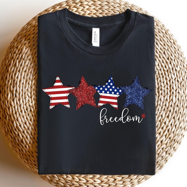 May include: Black t-shirt with three star designs in red, white, and blue, with a glittery texture. The word "freedom" is written in cursive below the stars. The shirt is made of a soft material.