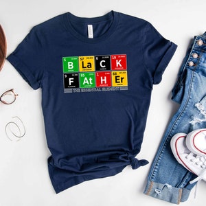 Op de afbeelding: Een marineblauw T-shirt met een afbeelding van het woord "BLACK" gespeld met behulp van chemische element symbolen. De afbeelding bevat ook de woorden "FATHER" gespeld met behulp van chemische element symbolen. De tekst "THE ESSENTIAL ELEMENT" is onder de afbeelding gedrukt.