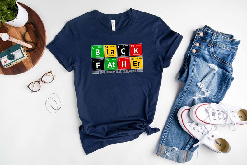 Op de afbeelding: Een marineblauw T-shirt met een afbeelding van het woord "BLACK" gespeld met behulp van chemische element symbolen. De afbeelding bevat ook de woorden "FATHER" gespeld met behulp van chemische element symbolen. De tekst "THE ESSENTIAL ELEMENT" is onder de afbeelding gedrukt.