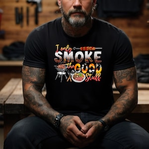 May include: A man wearing a black t-shirt with the text "I only smoke the good stuff" in orange and yellow lettering. The shirt features a grill, a plate of food, and a skewer of meat.