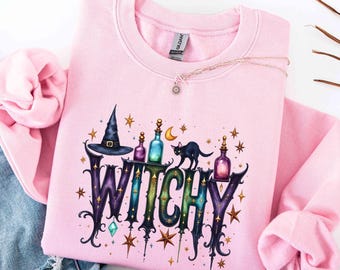 May include: A light pink crewneck sweatshirt featuring a graphic print of the word WITCHY in a stylized font. The design includes a black witch hat, a black cat, colorful potion bottles, a crescent moon, and gold stars scattered throughout.