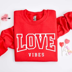 May include: Red sweatshirt with white text that says "LOVE VIBES" in a bold font. The sweatshirt is folded in half and there are heart-shaped candies and lollipops on a white background.