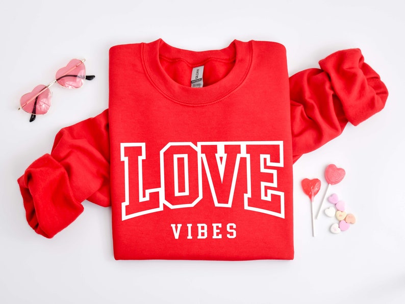 May include: Red sweatshirt with white text that says "LOVE VIBES" in a bold font. The sweatshirt is folded in half and there are heart-shaped candies and lollipops on a white background.