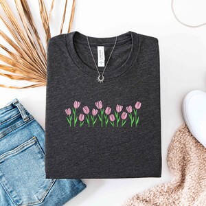 K&ouml;nnte beinhalten: Dunkelgraues T-Shirt mit einer gestickten Reihe rosa Tulpen mit gr&uuml;nen Stielen und Bl&auml;ttern. Eine silberne Halskette mit einem Sonnenanh&auml;nger liegt auf dem Shirt. Das Shirt wird mit blauen Jeans und wei&szlig;en Slip-On-Schuhen kombiniert.