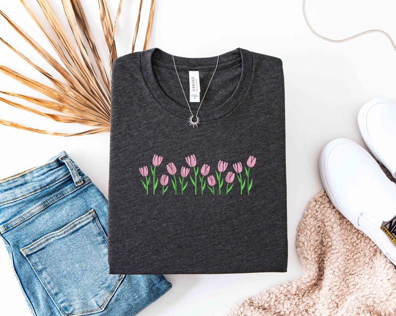 K&ouml;nnte beinhalten: Dunkelgraues T-Shirt mit einer gestickten Reihe rosa Tulpen mit gr&uuml;nen Stielen und Bl&auml;ttern. Eine silberne Halskette mit einem Sonnenanh&auml;nger liegt auf dem Shirt. Das Shirt wird mit blauen Jeans und wei&szlig;en Slip-On-Schuhen kombiniert.
