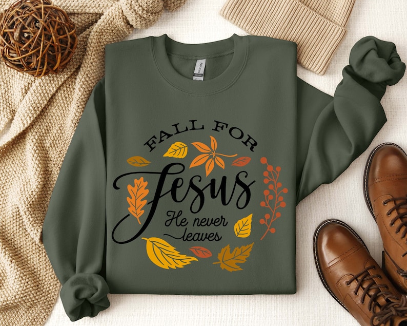 May include: A green sweatshirt with the text "Fall for Jesus He never leaves" in black lettering. The text is surrounded by a wreath of orange, yellow, and red leaves.
