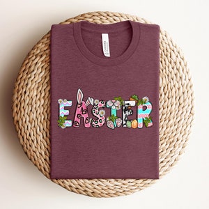 May include: A maroon t-shirt with the word "Easter" in colorful letters decorated with bunny ears, flowers, and other Easter-themed designs.