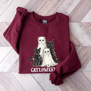 K&ouml;nnte beinhalten: Ein weinroter Pullover mit einem Aufdruck von drei schwarzen Katzen und zwei Geistern. Der Text "Catlowween" ist unter dem Aufdruck gedruckt.