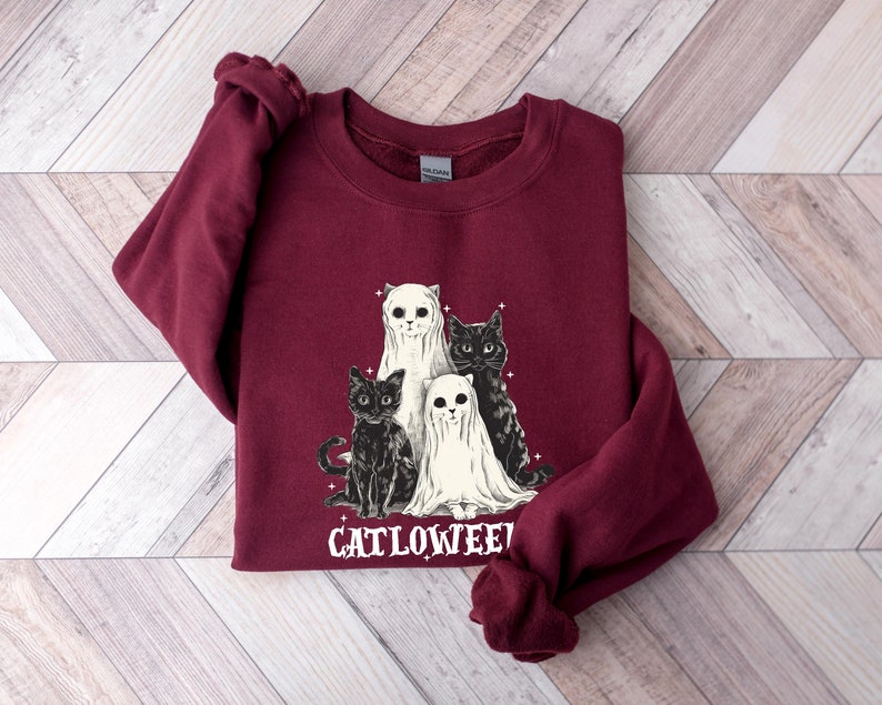 K&ouml;nnte beinhalten: Ein weinroter Pullover mit einem Aufdruck von drei schwarzen Katzen und zwei Geistern. Der Text "Catlowween" ist unter dem Aufdruck gedruckt.
