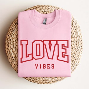 May include: Pink sweatshirt with the text "LOVE VIBES" in red block letters.