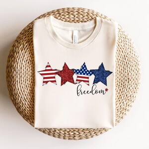 May include: A cream-coloured t-shirt with four star graphics in red, glittery red, blue with white stars, and dark blue glitter. The word "freedom" is written in a cursive font below the stars. The shirt is folded on a woven surface.
