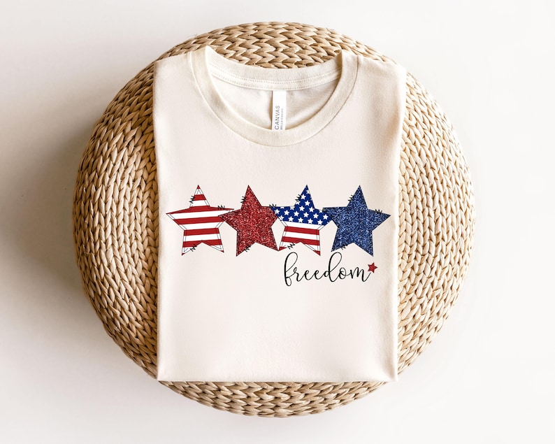 May include: A cream-coloured t-shirt with four star graphics in red, glittery red, blue with white stars, and dark blue glitter. The word "freedom" is written in a cursive font below the stars. The shirt is folded on a woven surface.