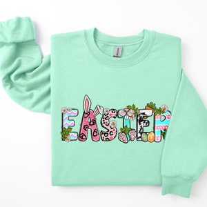 May include: A light green sweatshirt with the word "Easter" printed on the front in colorful letters. The letters are decorated with bunny ears, paw prints, and other Easter-themed designs.
