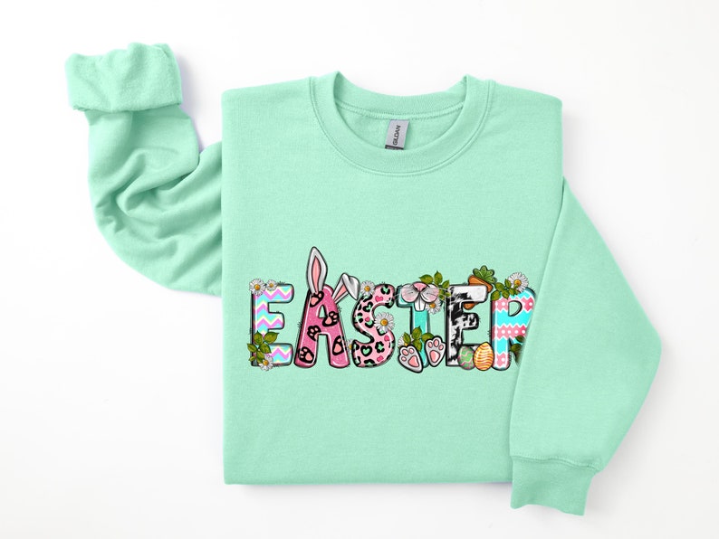 May include: A light green sweatshirt with the word "Easter" printed on the front in colorful letters. The letters are decorated with bunny ears, paw prints, and other Easter-themed designs.