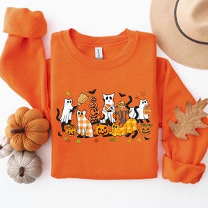 Może przedstawiać: Pomarańczowa bluza z nadrukiem graficznym kot&oacute;w przebranych na Halloween. Koty noszą kostiumy i trzymają dynie i słodycze. Bluza ma okrągły dekolt i długie rękawy.