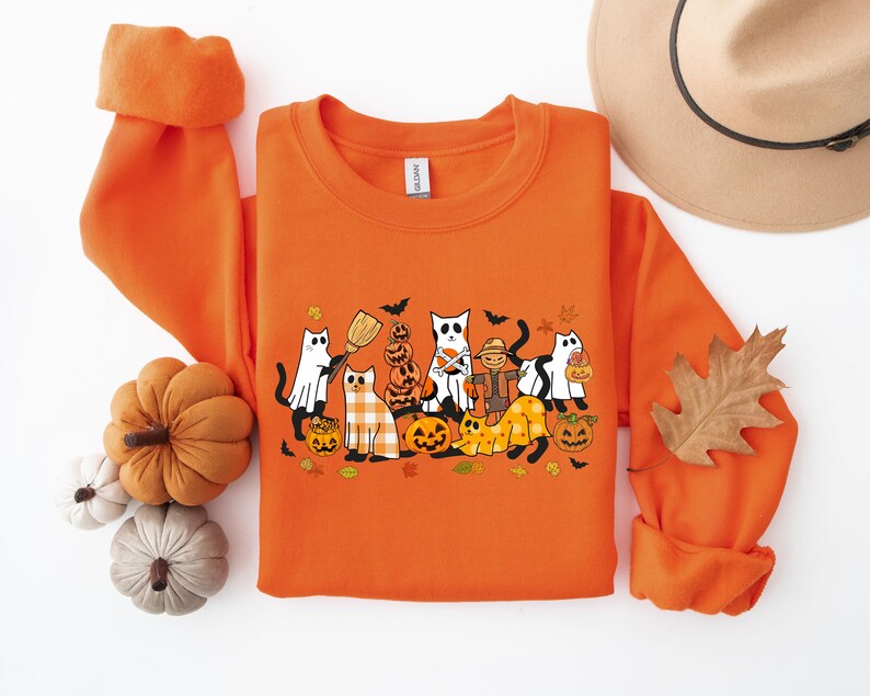 Może przedstawiać: Pomarańczowa bluza z nadrukiem graficznym kot&oacute;w przebranych na Halloween. Koty noszą kostiumy i trzymają dynie i słodycze. Bluza ma okrągły dekolt i długie rękawy.