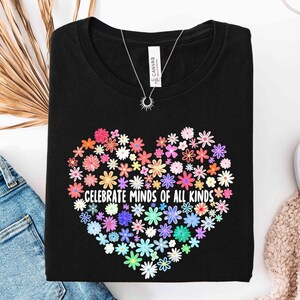 May include: A black t-shirt featuring a heart-shaped graphic composed of various colorful flowers. The text "CELEBRATE MINDS OF ALL KINDS" is printed in white across the center of the floral heart design. The shirt has a crew neckline.