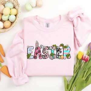 May include: Pink sweatshirt with the word "Easter" in colorful letters decorated with bunny ears, flowers, and eggs.