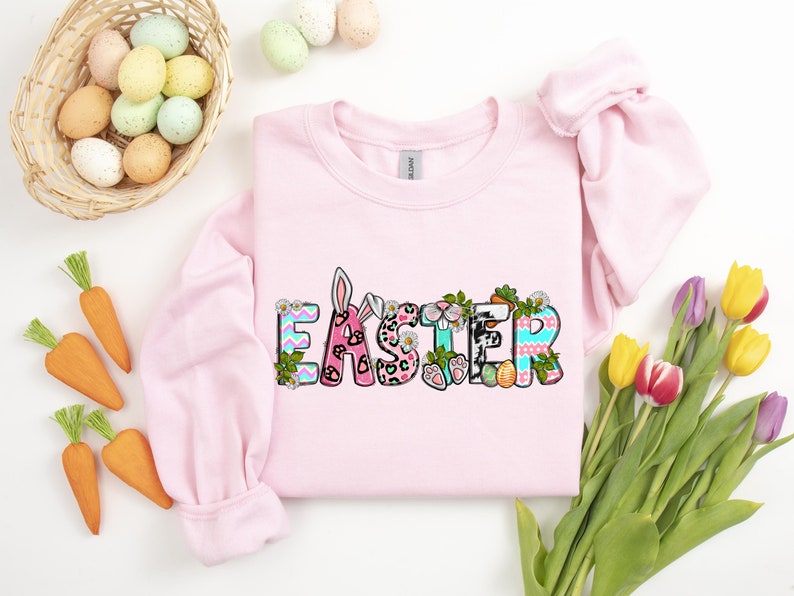 May include: Pink sweatshirt with the word "Easter" in colorful letters decorated with bunny ears, flowers, and eggs.