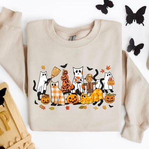 Może przedstawiać: Beżowa bluza z nadrukiem o tematyce Halloween przedstawiającym koty przebrane w kostiumy. Wz&oacute;r zawiera kota przebranego za ducha, kota przebranego za straszak, kota przebranego za czarownicę i kota przebranego za dynię. Wz&oacute;r zawiera r&oacute;wnież dynie, nietoperze i liście.