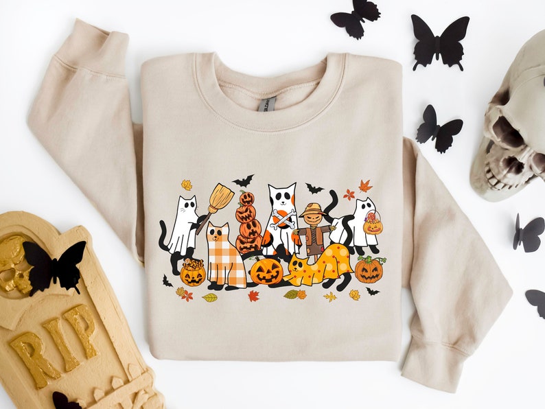 Może przedstawiać: Beżowa bluza z nadrukiem o tematyce Halloween przedstawiającym koty przebrane w kostiumy. Wz&oacute;r zawiera kota przebranego za ducha, kota przebranego za straszak, kota przebranego za czarownicę i kota przebranego za dynię. Wz&oacute;r zawiera r&oacute;wnież dynie, nietoperze i liście.