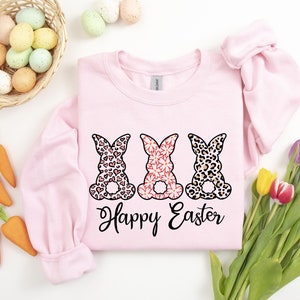 Puede incluir: Sudadera rosa con tres conejos en diferentes dise&ntilde;os estampados, corazones, flores e estampado de leopardo. Los conejos est&aacute;n sentados de espaldas al espectador. El texto "Happy Easter" est&aacute; impreso debajo de los conejos.
