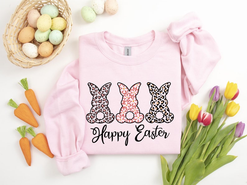 Puede incluir: Sudadera rosa con tres conejos en diferentes dise&ntilde;os estampados, corazones, flores e estampado de leopardo. Los conejos est&aacute;n sentados de espaldas al espectador. El texto "Happy Easter" est&aacute; impreso debajo de los conejos.