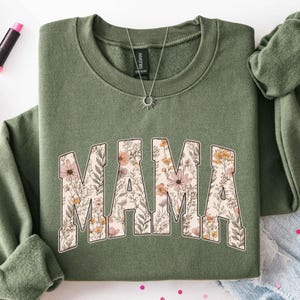 May include: An olive green sweatshirt with the word "MAMA" in large, floral-filled letters. The floral design includes pink, peach, and yellow flowers with green leaves. A silver necklace with a sunburst pendant is visible.