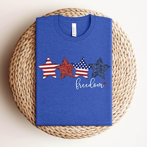 May include: A blue t-shirt with three star designs in red, glittery red, and navy blue with white stripes and stars. The word "freedom" is written in cursive below the stars. The shirt is displayed on a woven, circular surface.
