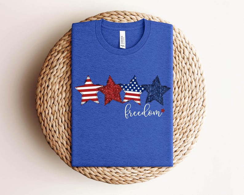 May include: A blue t-shirt with three star designs in red, glittery red, and navy blue with white stripes and stars. The word "freedom" is written in cursive below the stars. The shirt is displayed on a woven, circular surface.