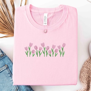 K&ouml;nnte beinhalten: Hellrosa Sweatshirt mit Rundhalsausschnitt und gestickten rosa Tulpen mit gr&uuml;nen Stielen und Bl&auml;ttern. Eine Halskette mit einem Sonnenanh&auml;nger ist sichtbar. Das Shirt liegt gefaltet auf einer wei&szlig;en Oberfl&auml;che.