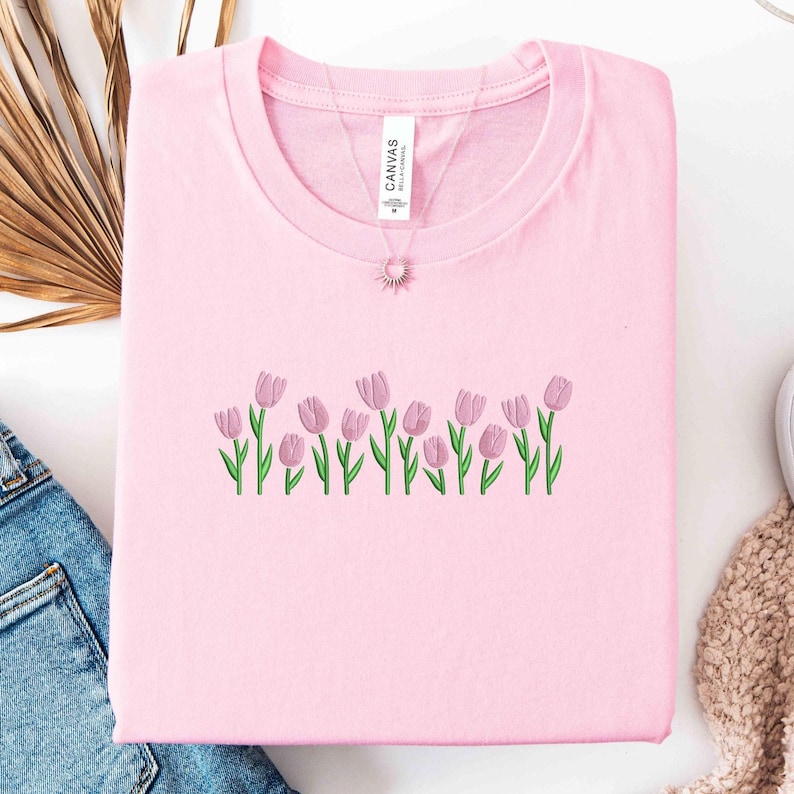K&ouml;nnte beinhalten: Hellrosa Sweatshirt mit Rundhalsausschnitt und gestickten rosa Tulpen mit gr&uuml;nen Stielen und Bl&auml;ttern. Eine Halskette mit einem Sonnenanh&auml;nger ist sichtbar. Das Shirt liegt gefaltet auf einer wei&szlig;en Oberfl&auml;che.