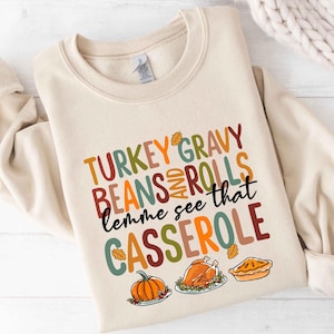 May include: Cream-colored sweatshirt with a Thanksgiving-themed graphic. The design features the words "TURKEY GRAVY BEANS AND ROLLS lemme see that CASSEROLE" in various colors, along with illustrations of a pumpkin, turkey, and pie.