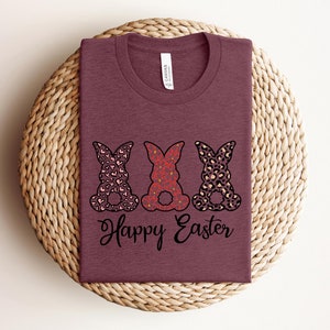 Puede incluir: Una camiseta color marr&oacute;n rojizo con tres siluetas de conejos en diferentes patrones. Los conejos llevan un patr&oacute;n de corazones rosa y blanco, un patr&oacute;n floral rojo y blanco y un estampado de leopardo marr&oacute;n y blanco. El texto "Happy Easter" est&aacute; impreso debajo de los conejos.