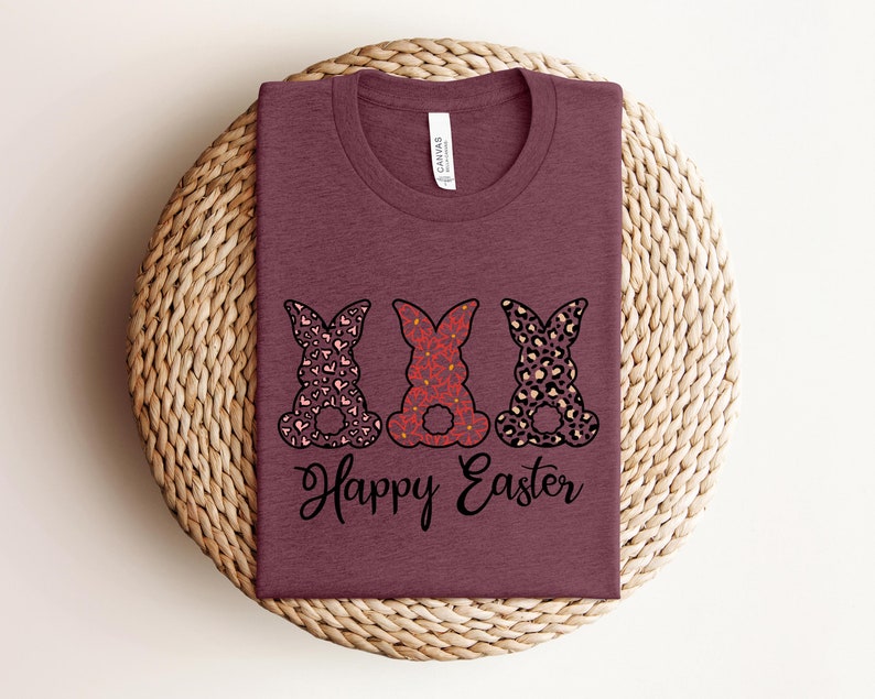 Puede incluir: Una camiseta color marr&oacute;n rojizo con tres siluetas de conejos en diferentes patrones. Los conejos llevan un patr&oacute;n de corazones rosa y blanco, un patr&oacute;n floral rojo y blanco y un estampado de leopardo marr&oacute;n y blanco. El texto "Happy Easter" est&aacute; impreso debajo de los conejos.