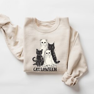 K&ouml;nnte beinhalten: Ein beiger Pullover mit einem schwarz-wei&szlig;en Motiv von drei Katzen, die als Geister verkleidet sind. Der Text "Catlowween" ist unter dem Motiv gedruckt.