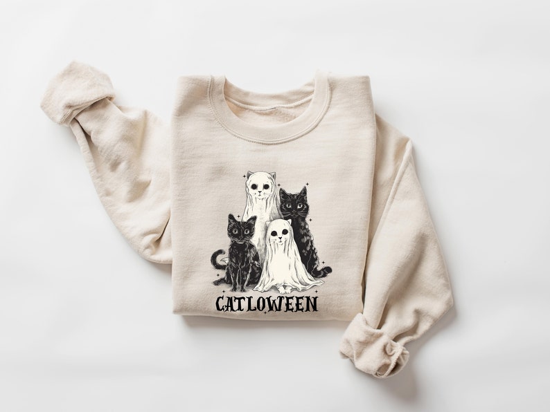 K&ouml;nnte beinhalten: Ein beiger Pullover mit einem schwarz-wei&szlig;en Motiv von drei Katzen, die als Geister verkleidet sind. Der Text "Catlowween" ist unter dem Motiv gedruckt.