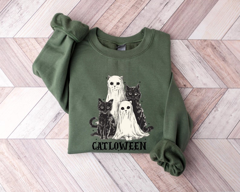 K&ouml;nnte beinhalten: Ein gr&uuml;ner Pullover mit einem schwarz-wei&szlig;en Motiv von drei Katzen, die als Geister verkleidet sind. Der Text "Catlloween" ist unter dem Motiv gedruckt.