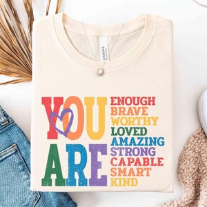 May include: Cream-colored t-shirt with the words "YOU ARE" in rainbow colors, with a purple heart graphic. Below, the words "ENOUGH, BRAVE, WORTHY, LOVED, AMAZING, STRONG, CAPABLE, SMART, KIND" are printed in various colors.
