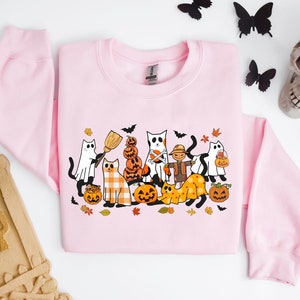 Może przedstawiać: R&oacute;żowa bluza z nadrukiem o tematyce Halloween przedstawiającym grupę kot&oacute;w przebranych w kostiumy i trzymających dynie. Koty noszą r&oacute;żne kostiumy, w tym ducha, czarownicę, straszak i dynię. Nadruk jest otoczony jesiennymi liśćmi i dyniami.