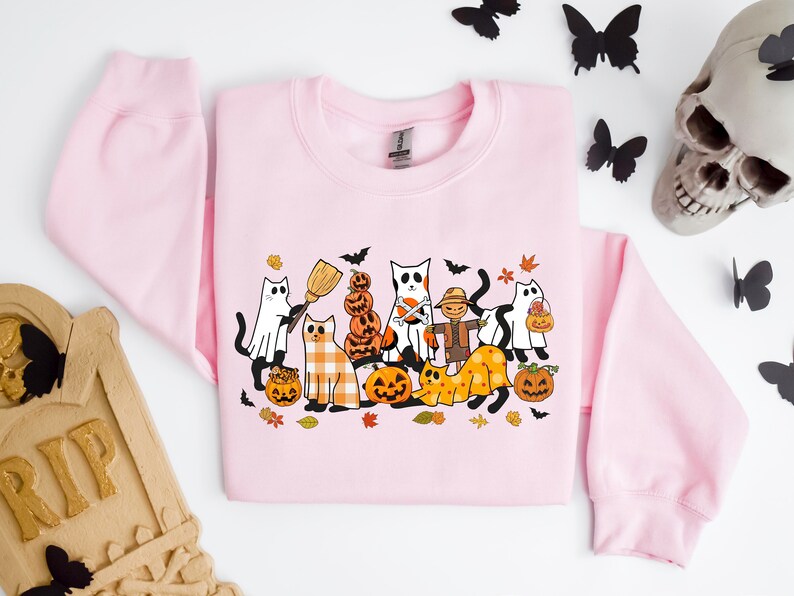 Może przedstawiać: R&oacute;żowa bluza z nadrukiem o tematyce Halloween przedstawiającym grupę kot&oacute;w przebranych w kostiumy i trzymających dynie. Koty noszą r&oacute;żne kostiumy, w tym ducha, czarownicę, straszak i dynię. Nadruk jest otoczony jesiennymi liśćmi i dyniami.