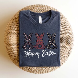 Puede incluir: Una camiseta gris oscuro con tres siluetas de conejos en diferentes dise&ntilde;os estampados. Los conejos est&aacute;n en fila y tienen el texto "Happy Easter" debajo.