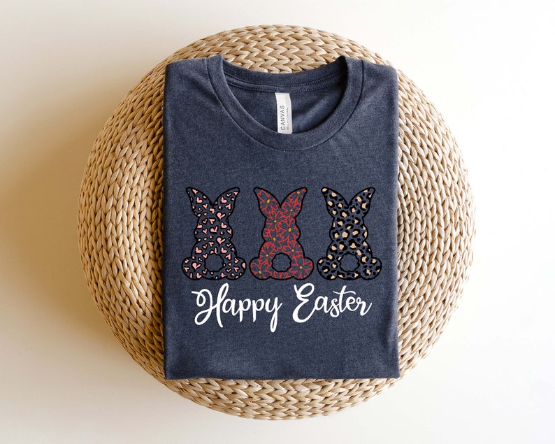 Puede incluir: Una camiseta gris oscuro con tres siluetas de conejos en diferentes dise&ntilde;os estampados. Los conejos est&aacute;n en fila y tienen el texto "Happy Easter" debajo.