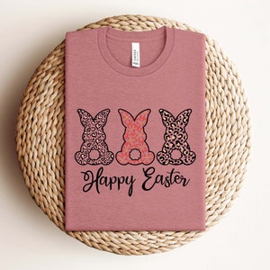 Puede incluir: Una camiseta color malva con tres siluetas de conejos. Los conejos est&aacute;n estampados con corazones, flores e impresi&oacute;n de leopardo. El texto "Happy Easter" est&aacute; impreso debajo de los conejos.