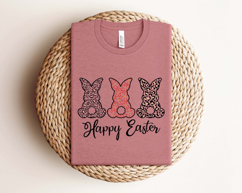 Puede incluir: Una camiseta color malva con tres siluetas de conejos. Los conejos est&aacute;n estampados con corazones, flores e impresi&oacute;n de leopardo. El texto "Happy Easter" est&aacute; impreso debajo de los conejos.